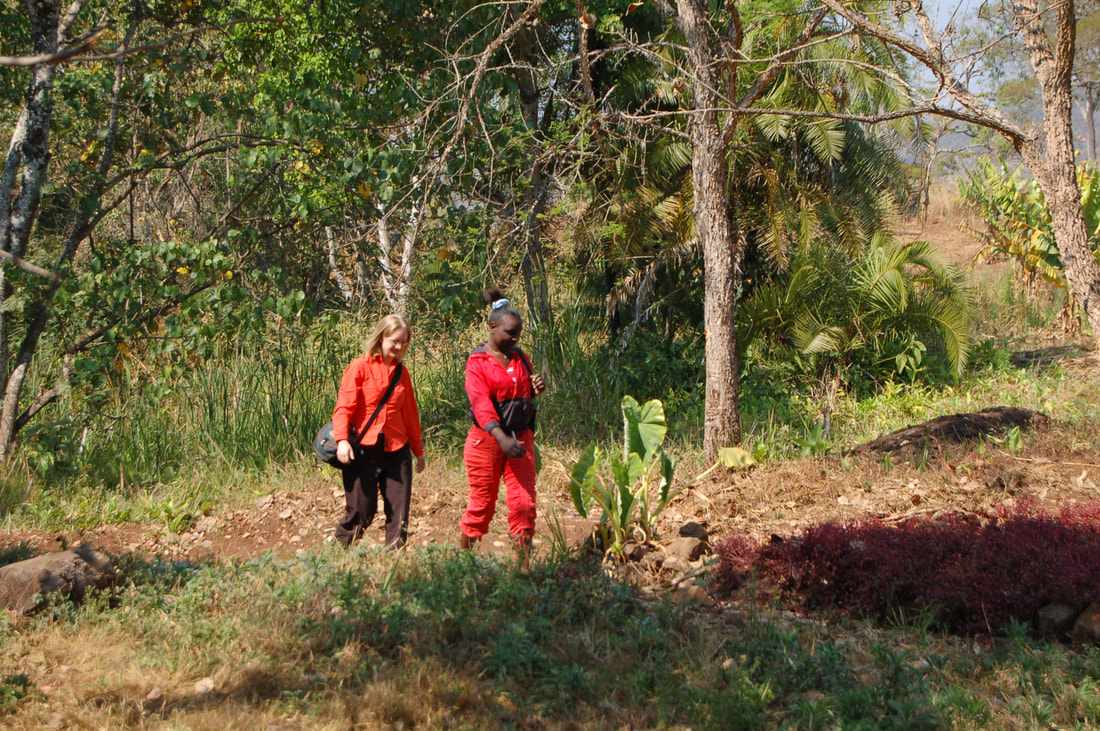 Two women walk on a path in a rural area with tropical trees in the background.