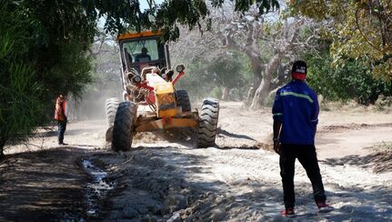 Two workers observe as a third man drives a large road grader over a damaged road.