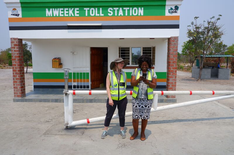 Two women stand next to a vehicle gate in front of a small toll building reading "Mweeke Toll Station." The women are looking at a mobile phone together and laughing.